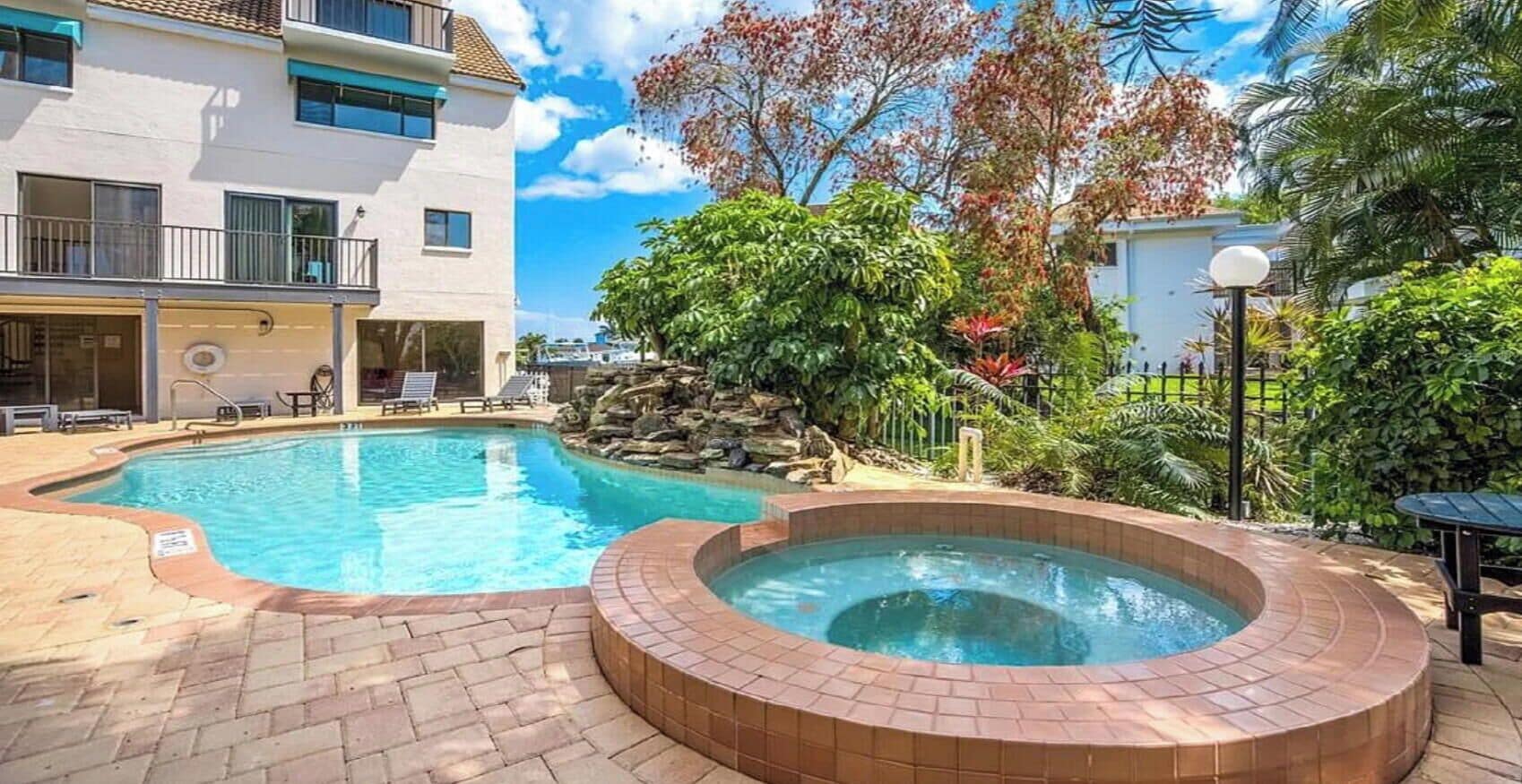 Une piscine avec un jacuzzi, entourée d’une terrasse en briques et d’une végétation luxuriante.