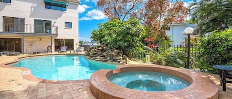 Una piscina con hidromasaje, rodeada de un patio de ladrillo y exuberante vegetación.