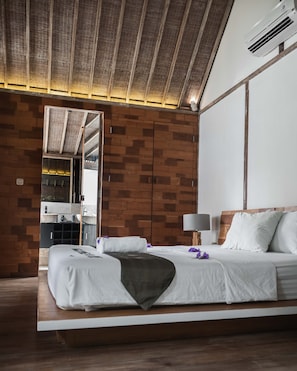 A modern hotel room with a wooden ceiling, a large bed with white linens, a bedside table with a lamp, and a bathroom visible through an open door.