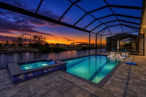 Una piscina con cerramiento de vidrio, un jacuzzi y una zona de estar cubierta con vistas al atardecer.