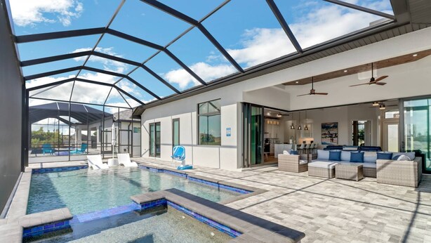 A modern indoor pool area with a glass ceiling, a seating area with sofas, and a kitchen visible through glass doors.