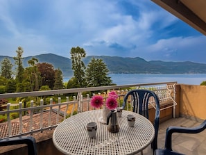 A balcony with a round table, two chairs, a vase with flowers, and a view of a lake and mountains.
