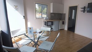 Una cocina moderna con armarios blancos, microondas y cafetera. Hay una mesa de vidrio con sillas y una puerta que da a otra habitación.