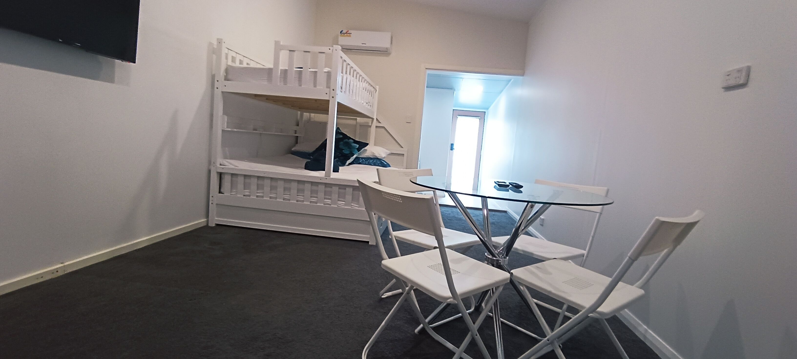 Een kamer met stapelbedden, een klein tafeltje met stoelen, en een deur naar een andere kamer.