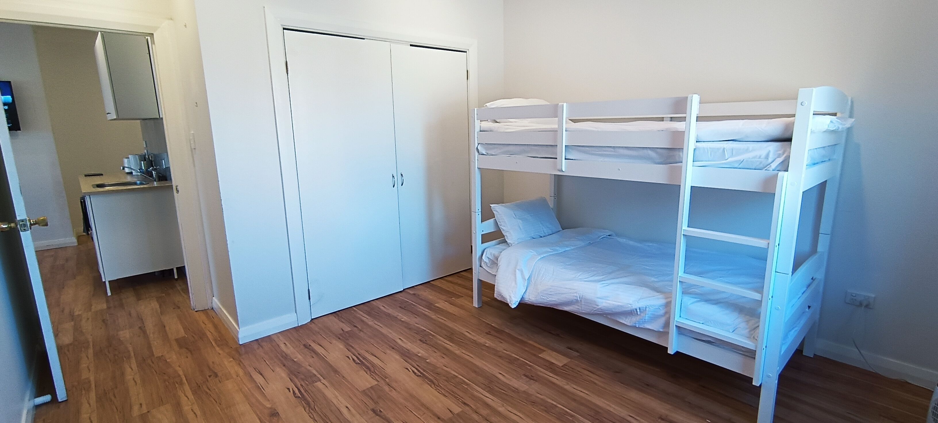 Ein Schlafraum mit Etagenbetten, Holzboden, einem weißen Kleiderschrank und einer kleinen Küchenzeile.