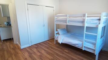 Ein Schlafraum mit Etagenbetten, Holzboden, einem weißen Kleiderschrank und einer kleinen Küchenzeile.