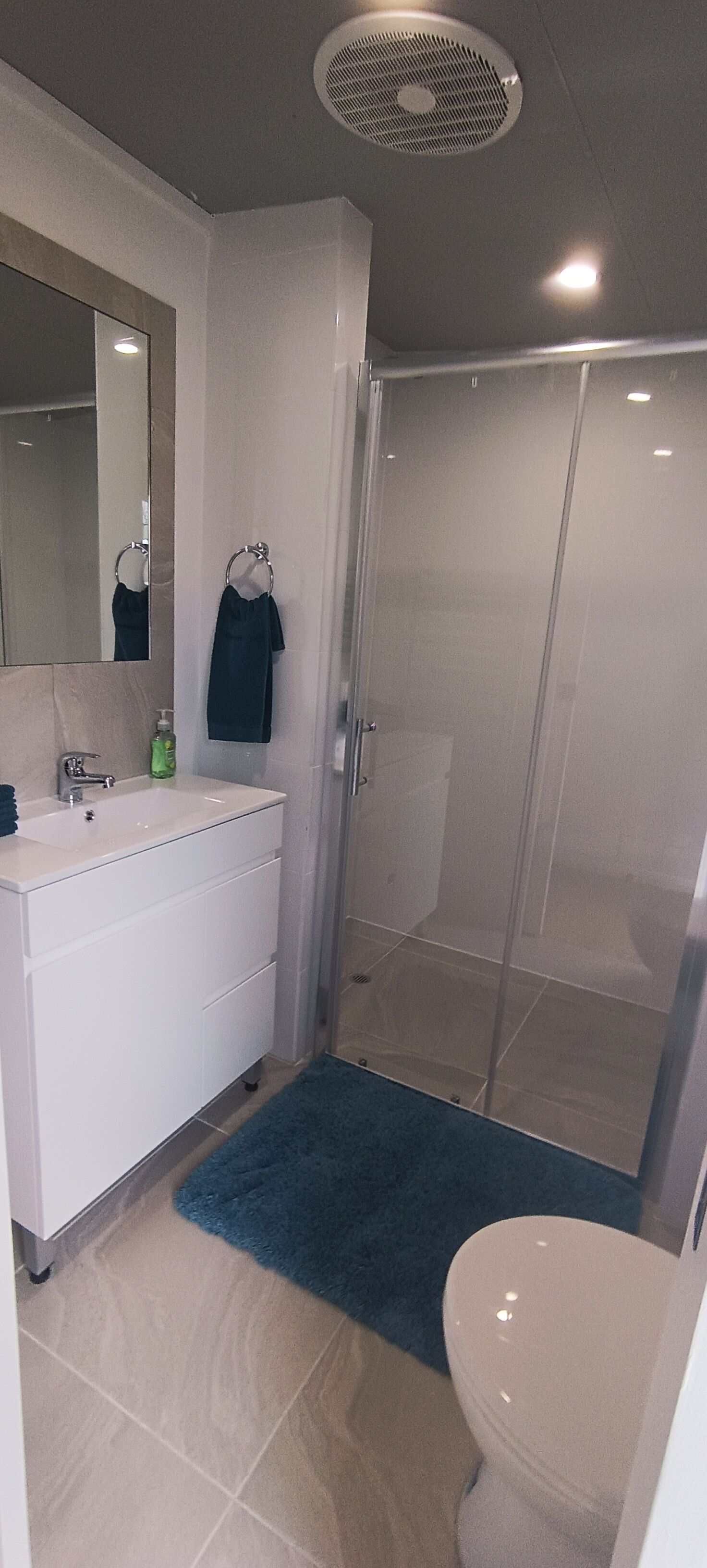 A modern bathroom with a white sink, mirror, and toilet. It includes a glass shower enclosure and a blue rug.