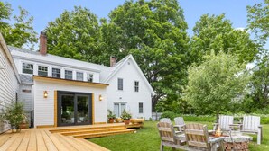 Une terrasse en bois avec un foyer et des fauteuils Adirondack, une maison blanche dotée d’une porte-fenêtre coulissante et une cheminée en briques.