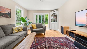 Un salon moderne avec un canapé gris, un fauteuil gris plus petit, une table basse en bois et un grand téléviseur posé sur un meuble en bois. Un tapis à motifs recouvre le sol, et une grande fenêtre offre une vue sur la verdure à l’extérieur.