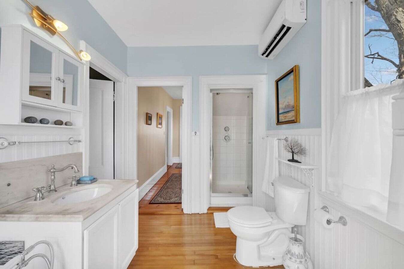 Une salle de bain avec un lavabo en marbre, des toilettes et une douche. Le sol est en bois et une porte blanche donne accès à une autre pièce.