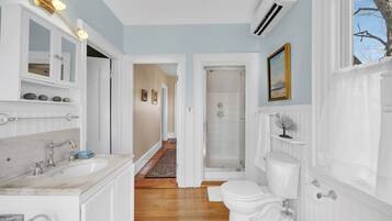 Une salle de bain avec un lavabo en marbre, des toilettes et une douche. Le sol est en bois et une porte blanche donne accès à une autre pièce.