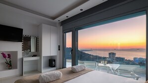 A modern bedroom with a large bed, a vanity with a mirror, and a view of the sunset over the water. - Rhodes Skyline Suite With out Door Jacuzzi Sea View A1 (Rhodes)