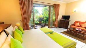 A hotel room with a large bed, a sofa, a TV, and a sliding glass door leading to a balcony with plants. - Ayurveda Spring of life Resort (Piliyandala)
