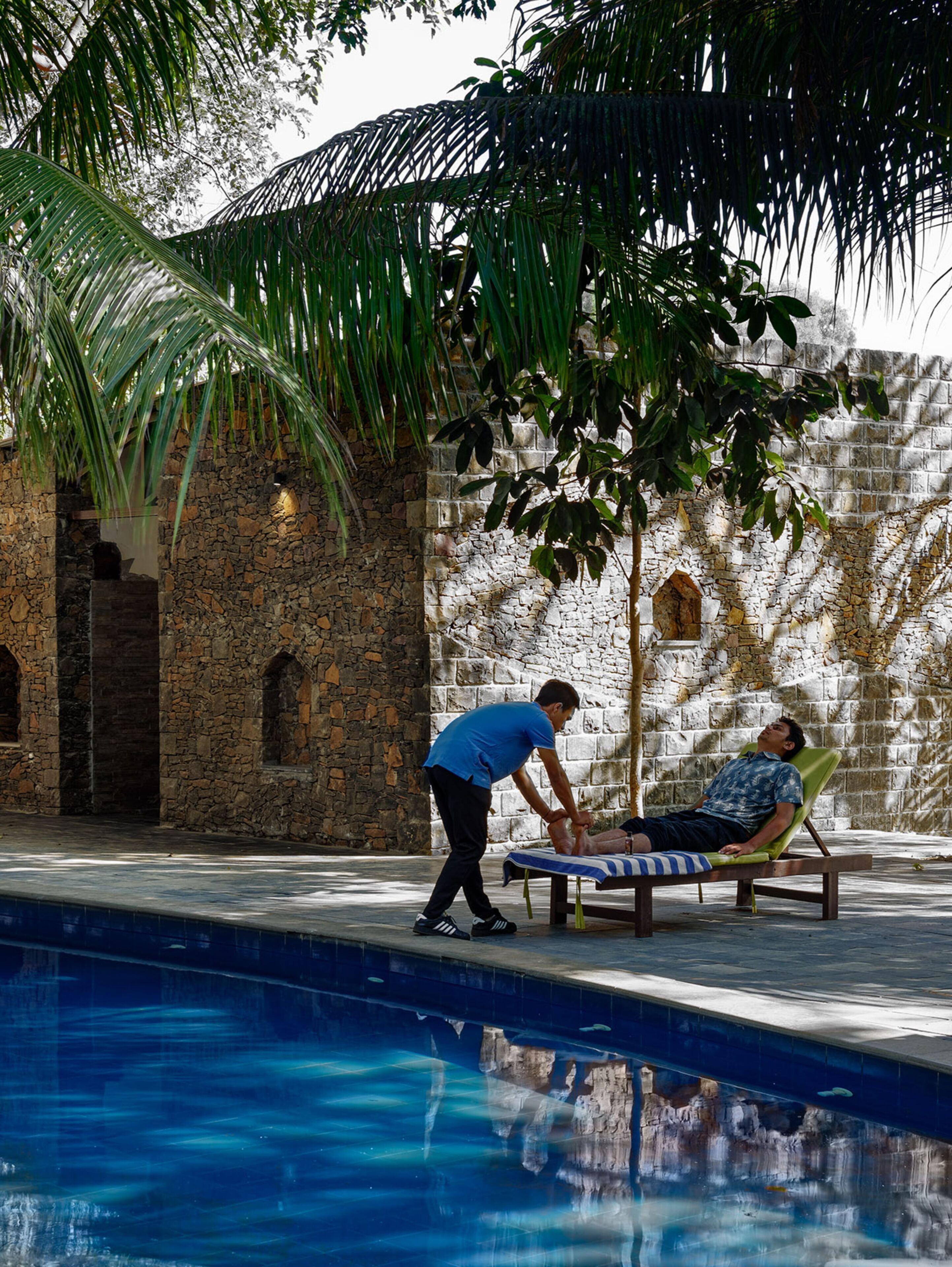 A pool area with a stone wall, a palm tree, and a person attending to someone lying on a lounge chair.
