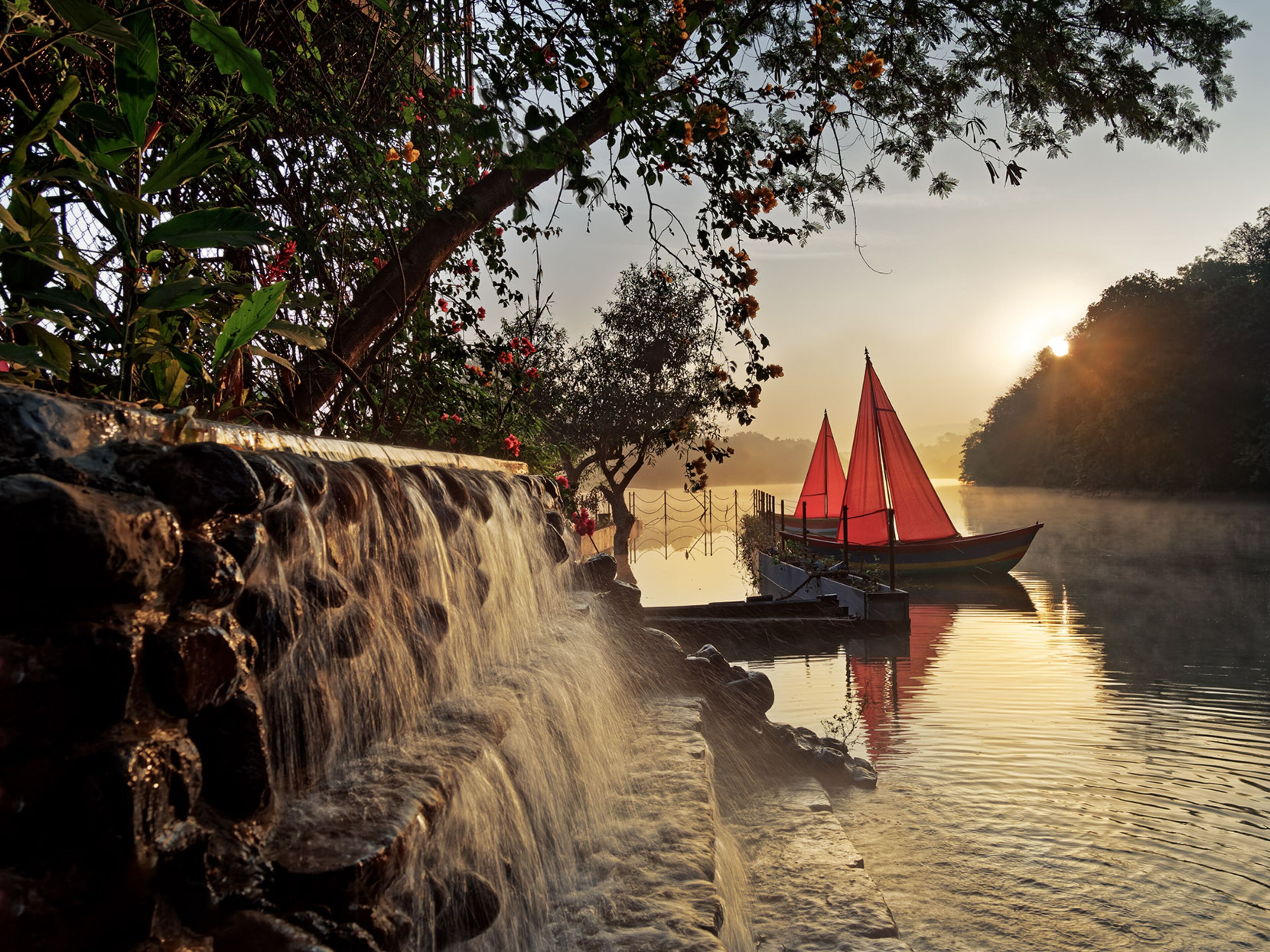Een waterval, een boot met rode zeilen en een mistig meer bij zonsopgang.