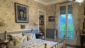 A bedroom with a floral-patterned bedspread, a wooden bed frame, a nightstand with a lamp, a bookshelf, and a window with curtains. - Champs Élysées (Paris)