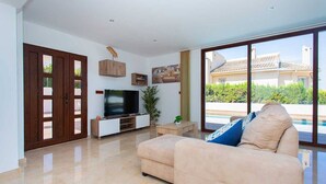 Un soggiorno moderno con una TV a schermo piatto, un divano color beige e un'ampia porta scorrevole in vetro che dà su una zona piscina.