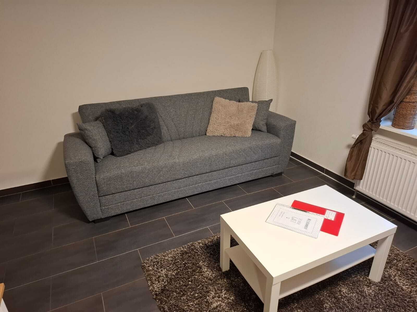 Ein graues Sofa mit zwei Kissen, ein weißer Tisch mit Unterlagen und ein brauner Teppich.