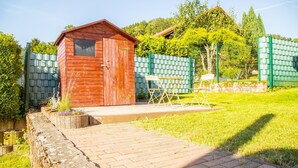 Eine Holzhütte mit Tür und Fenster, einem kleinen Tisch und Stuhl, sowie einem Pflasterweg, der zu einem grünen Zaun führt.