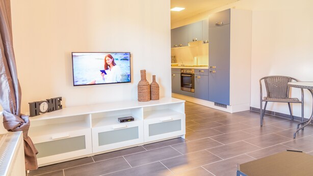 Ein modernes Wohnzimmer mit einem Flachbildfernseher, einer weißen Unterhaltungseinheit, Korbstühlen und einem Küchenbereich im Hintergrund.