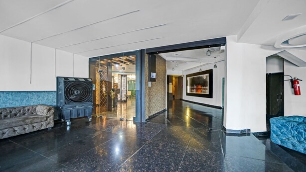 A modern lobby with a black-tiled floor, a blue upholstered sofa, a large fan, and a wall-mounted fire extinguisher. - The Bagga Inn (Jabalpur)