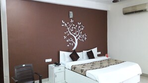 A bedroom with a bed, white bedding, a brown wall with a tree decal, a desk with a chair, and a red object on the desk. - The Bagga Inn (Jabalpur)