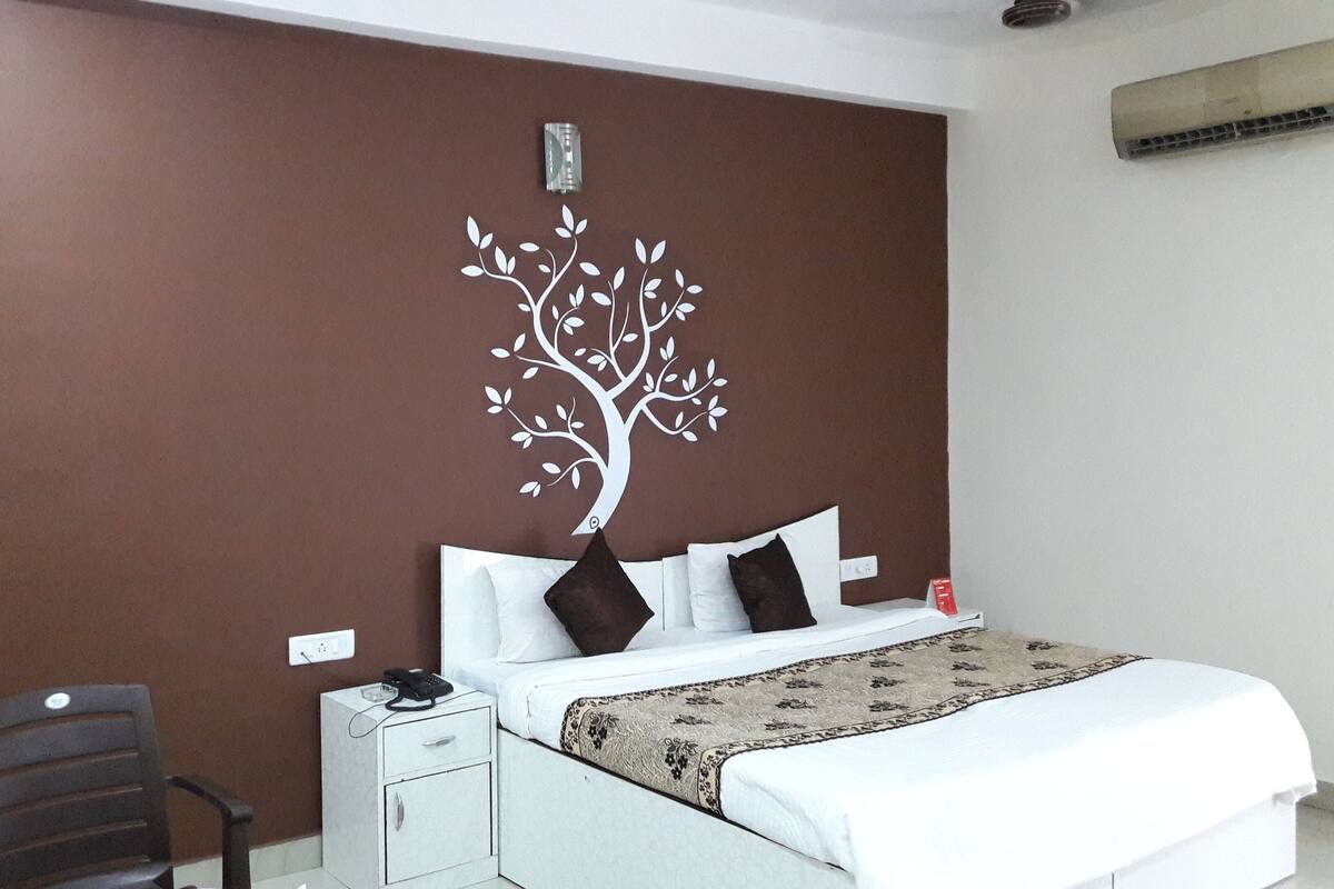 A bedroom with a bed, white bedding, a brown wall with a tree decal, a desk with a chair, and a red object on the desk.