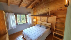 Quarto de cabana de madeira com cama, cadeira suspensa, janela com cortinas e um abajur.
