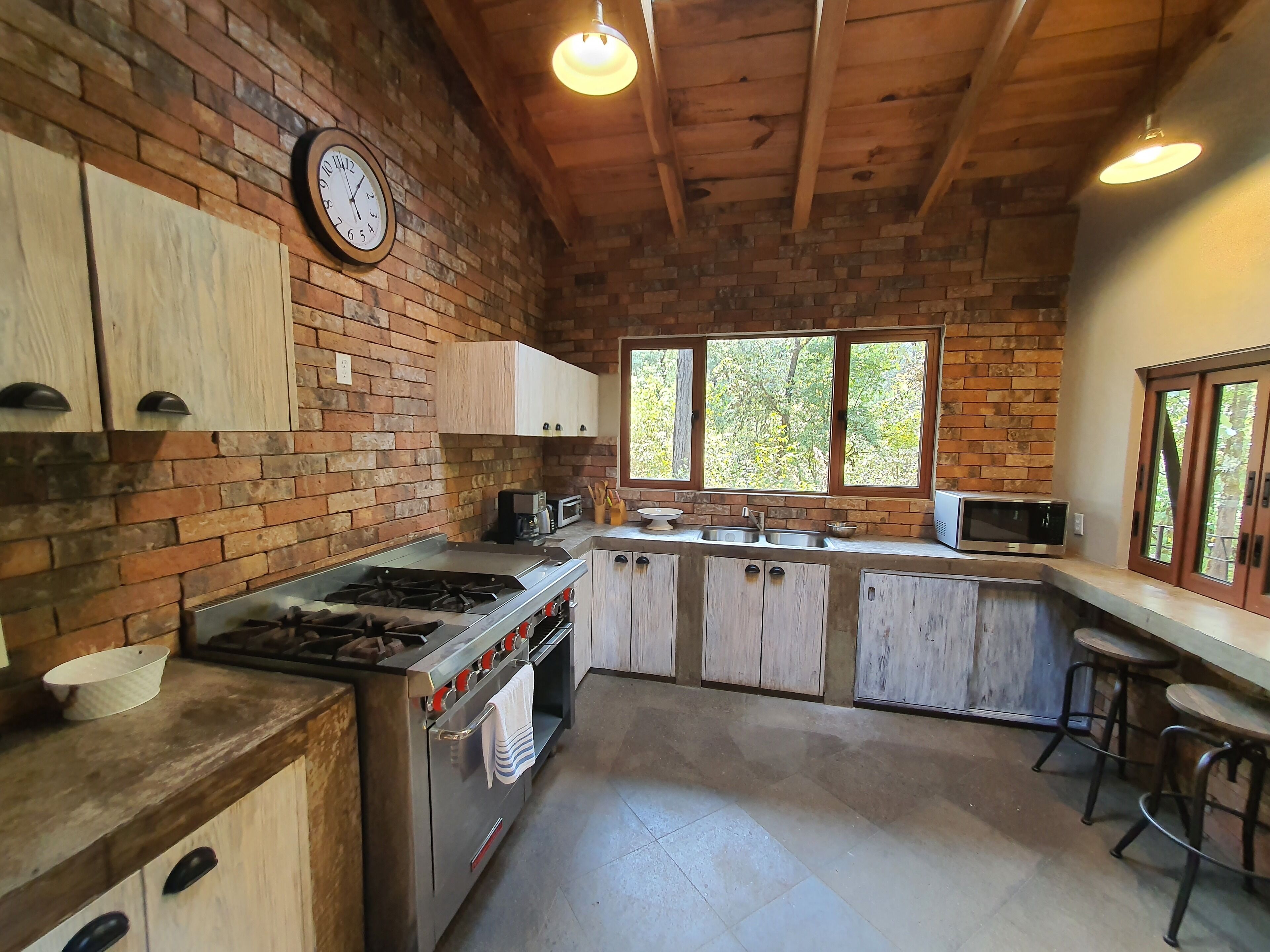 A rustic kitchen with a brick wall, wooden cabinets, a gas stove, a microwave, and a window overlooking greenery.