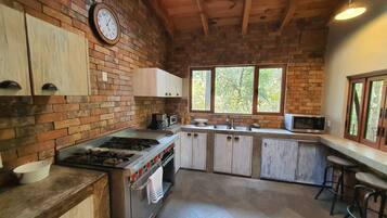 A rustic kitchen with a brick wall, wooden cabinets, a gas stove, a microwave, and a window overlooking greenery.