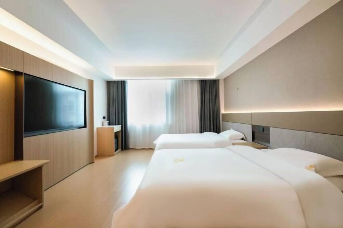 A modern hotel room with a flat-screen TV, two beds, a bedside table, and a large window with curtains.