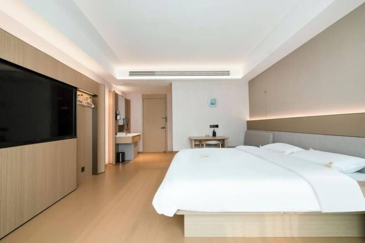 A modern hotel room with a large bed, a flat-screen TV, a bedside table, and a bathroom with a sink and mirror.