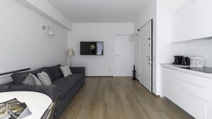 A modern, minimalist living space with a grey sofa, a flat-screen TV, a dining table, and a kitchen area with white cabinets and a coffee machine. - Italianway Collection - Corso Como 11 - C (Milan)