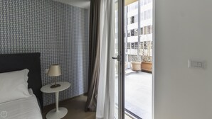 Ett hotellrum med tapet med ett mönster, en säng, ett runt bord med en lampa och en skjutdörr i glas som leder ut till ett utomhusområde med krukväxter.
