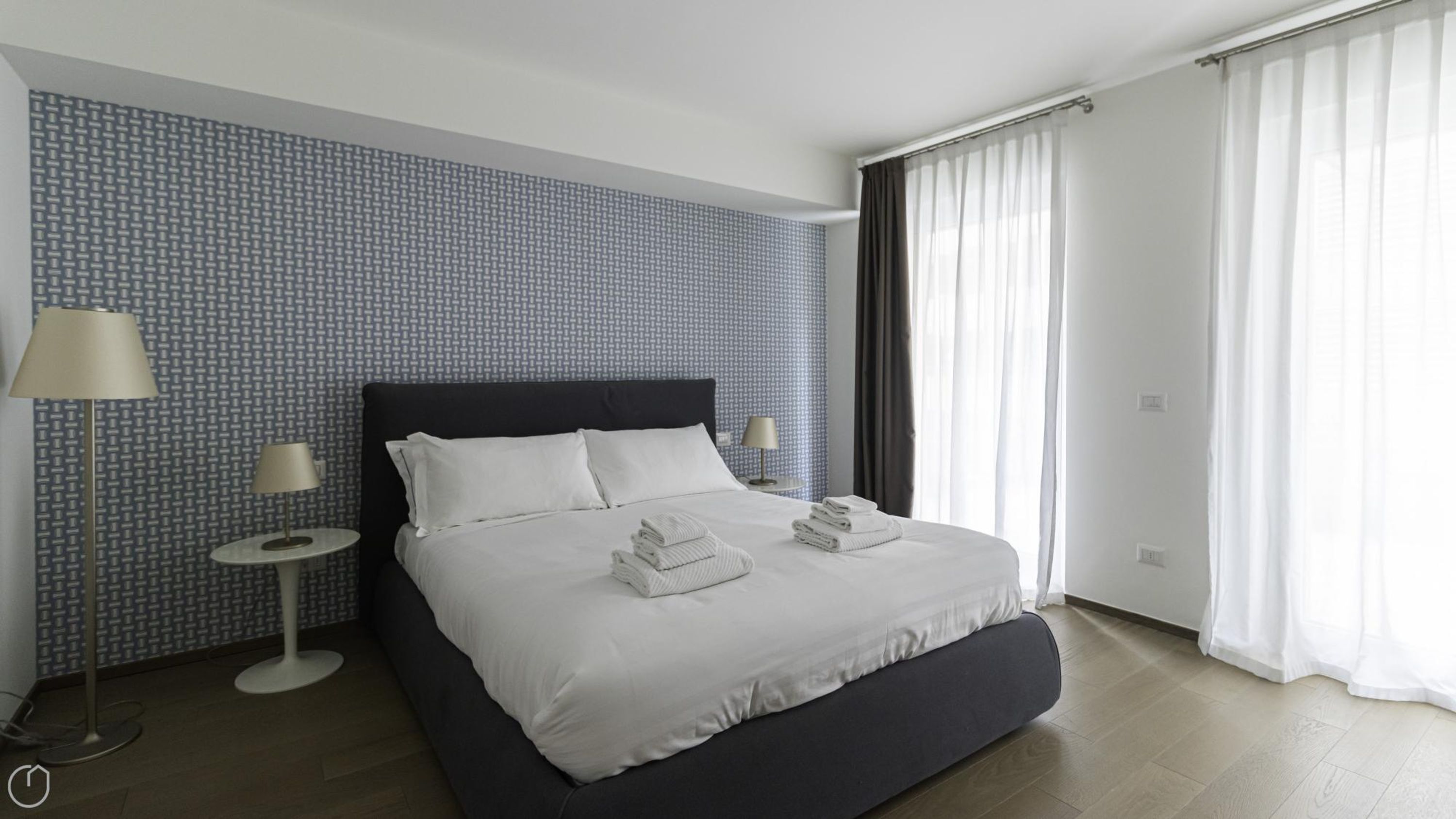 A modern hotel room with a large bed, two bedside tables, and a view of the outdoors through sheer curtains.