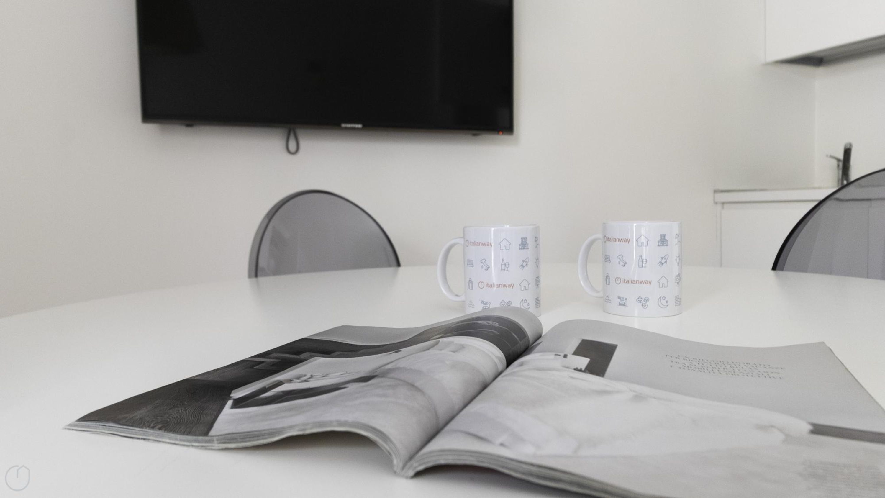 A modern meeting room with a flat-screen TV mounted on the wall, two white mugs with Italianway branding, and a magazine on the table.