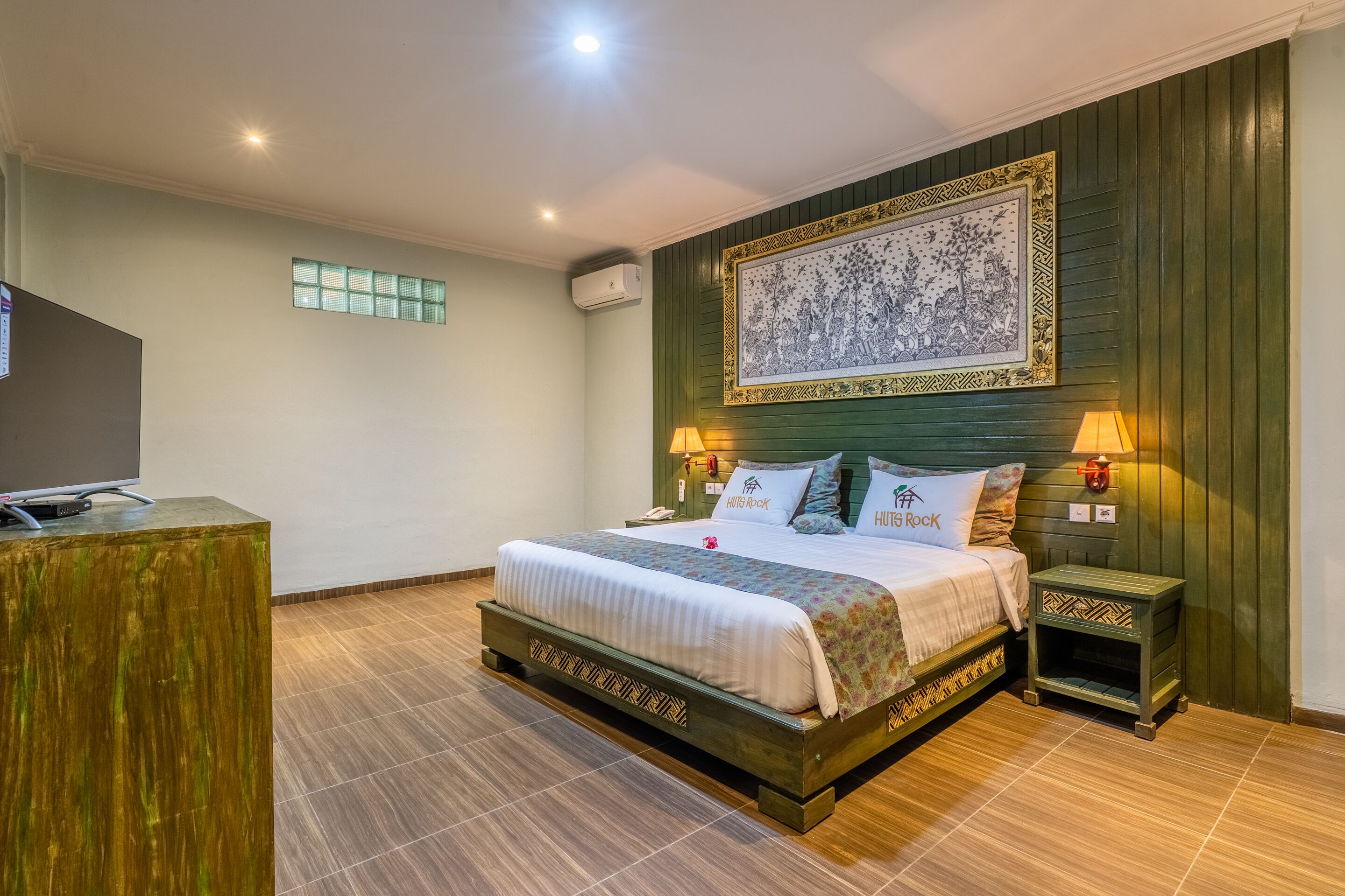 A hotel room with a large bed, two bedside tables with lamps, a flat-screen TV, a decorative wall hanging, and a small side table.