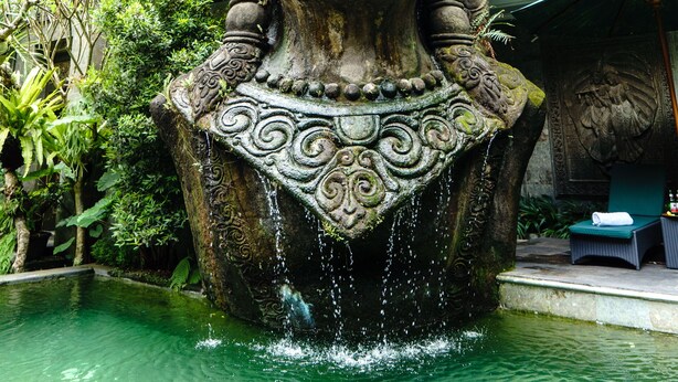 Eine große Steinstatue mit aufwendigen Schnitzereien, die über einem Wasserspiel in einem Innenhof mit üppiger Bepflanzung und einem Gebäude im Hintergrund platziert ist.