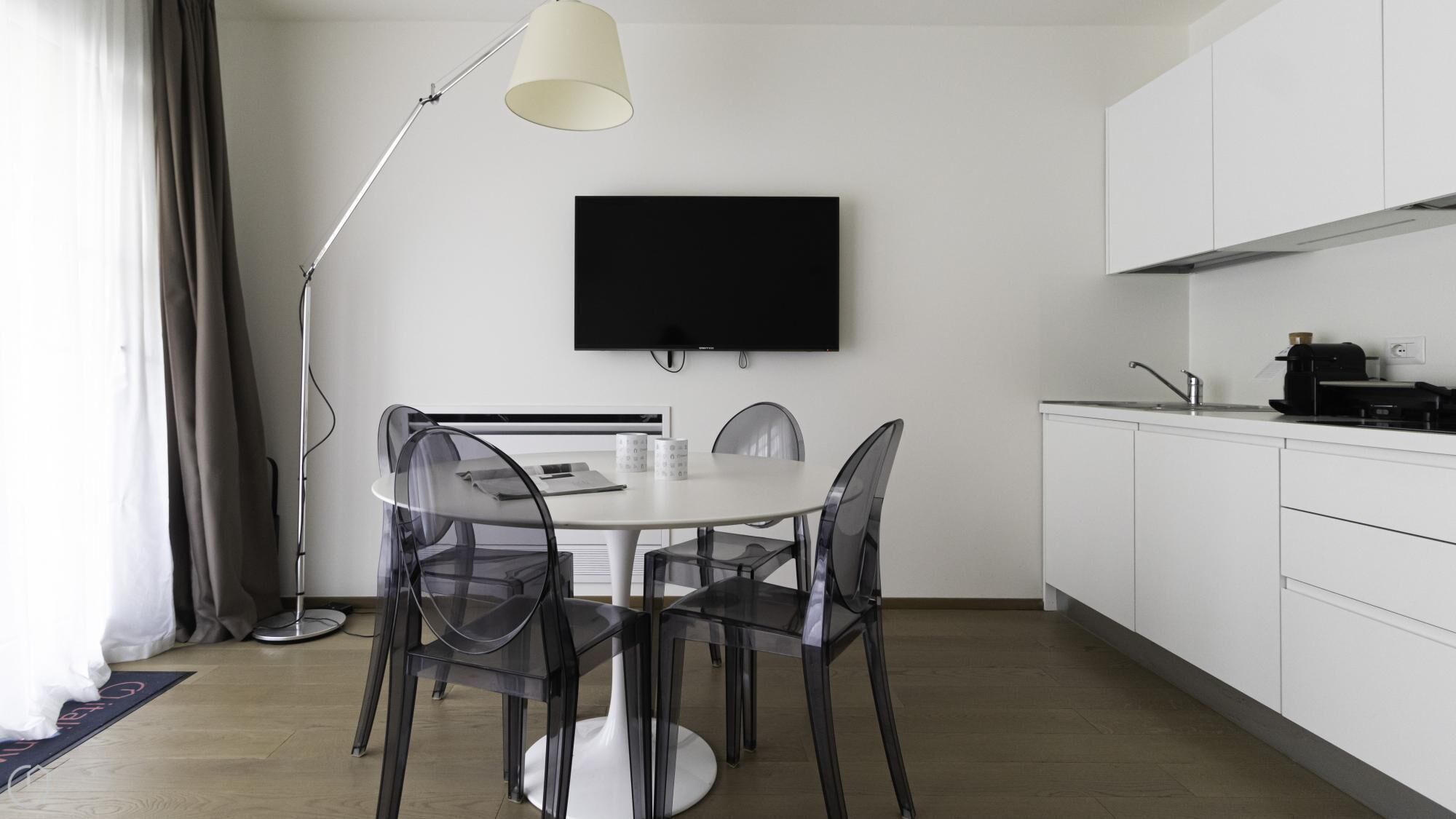 A modern kitchen with a dining area, white cabinets, a flat-screen TV, and a pendant light.