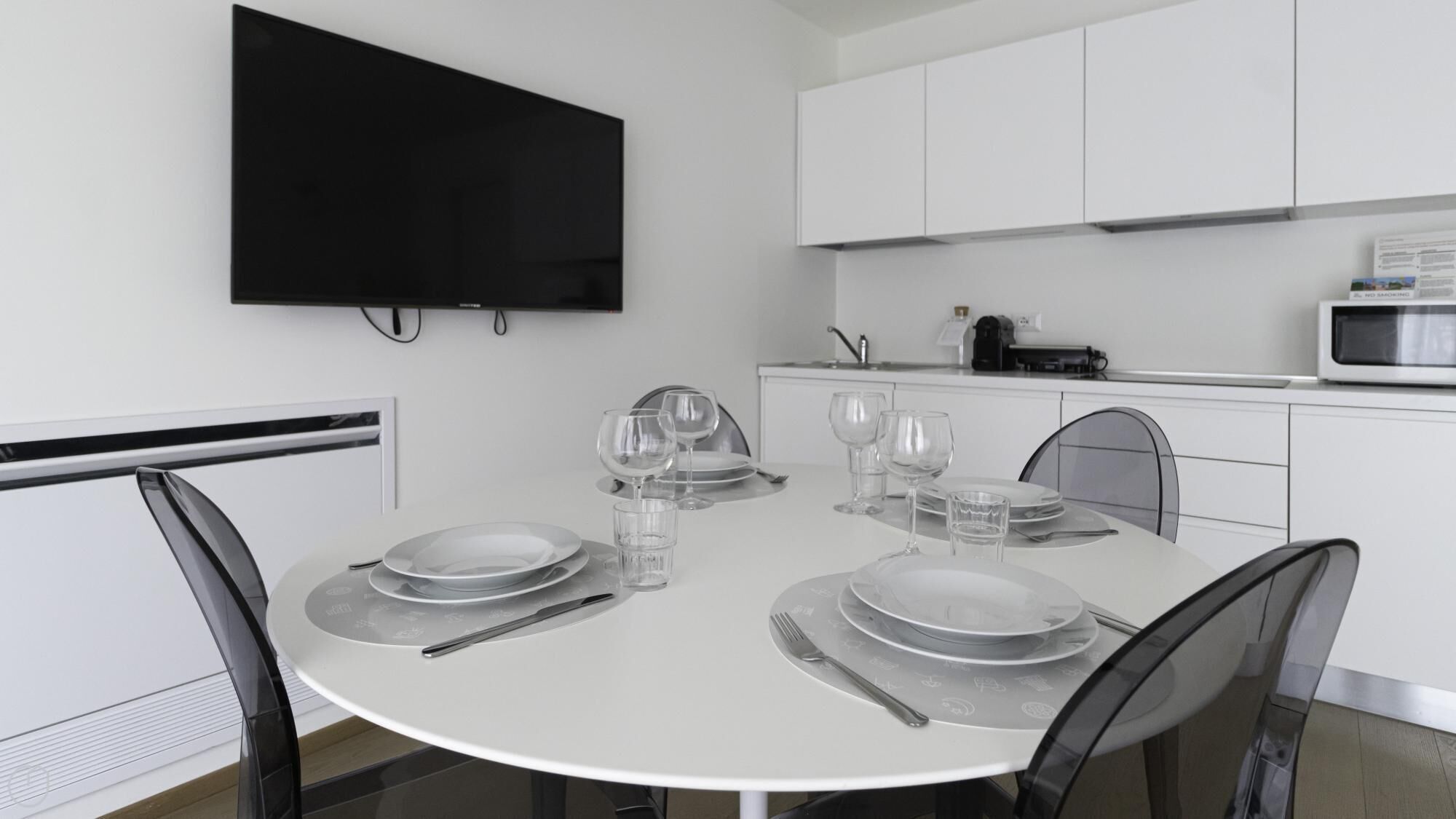 A modern kitchen with white cabinets, a flat-screen TV mounted above the countertop, and a dining table set with plates, glasses, and cutlery.