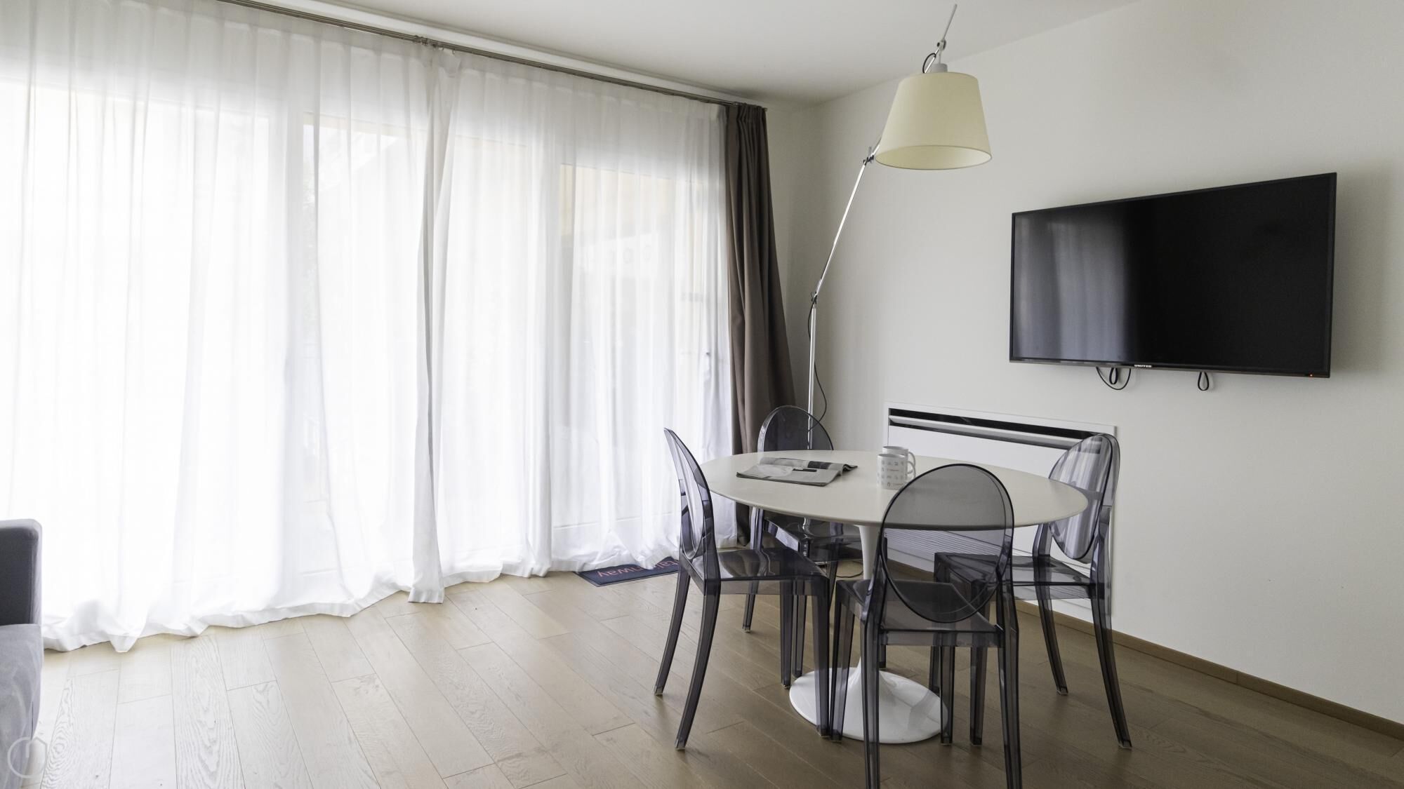 A modern dining area with a round table, clear chairs, a floor lamp, and a flat-screen TV mounted on the wall.