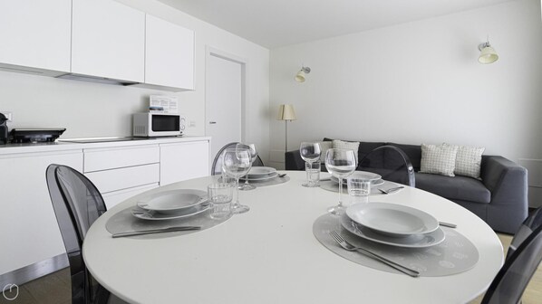 Un comedor moderno y minimalista con una mesa redonda para cuatro personas, armarios blancos, un microondas y un sofá al fondo.
