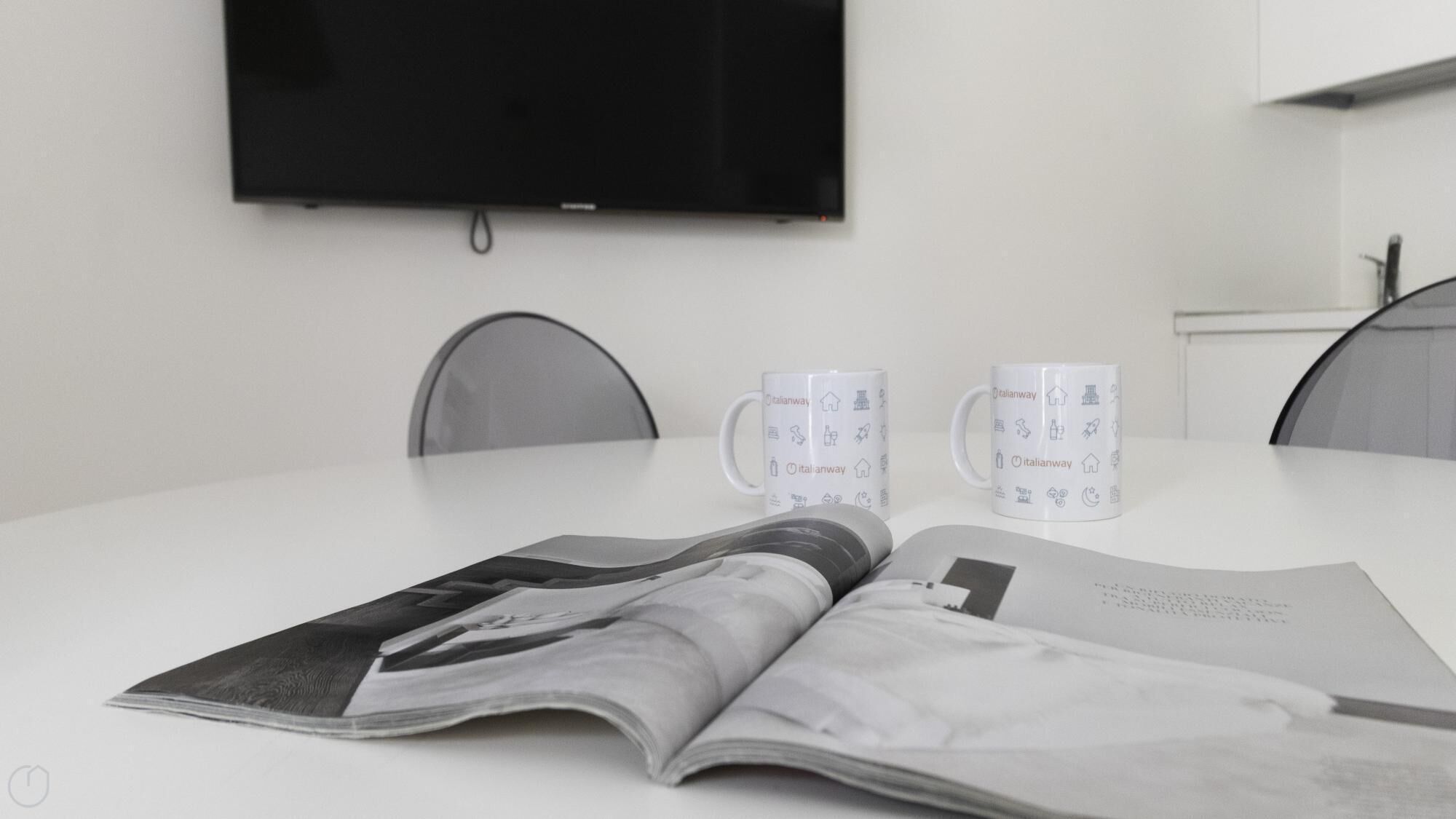 Una habitación moderna y minimalista con un televisor de pantalla plana en la pared, dos tazas blancas con motivos italianos y una revista sobre la mesa.