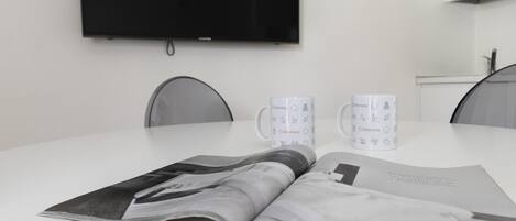 Una habitación moderna y minimalista con un televisor de pantalla plana en la pared, dos tazas blancas con motivos italianos y una revista sobre la mesa.