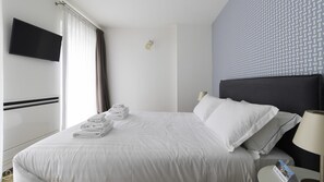 Habitación de hotel con cama, televisión montada en la pared, ventana con cortinas, lámpara de noche y pared con diseño.