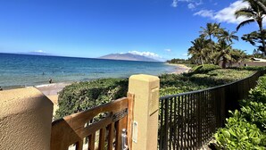 A beach view with a wooden fence, palm trees, and a mountain in the distance. - Ekahi 27A: Priceless Ocean View from a Private Lanai (Kihei)