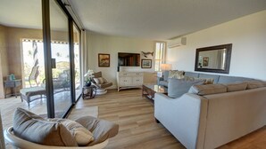 A spacious living area with a sofa, a dining table, and a kitchen area visible through a glass door. - Ekahi 27A: Priceless Ocean View from a Private Lanai (Kihei)