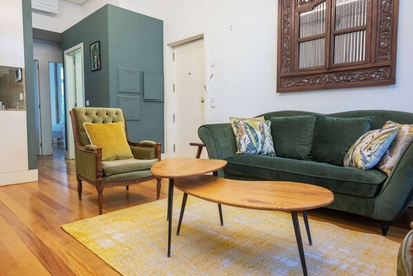Un salon avec un canapé vert, un fauteuil en bois avec un coussin jaune et une table basse ronde en bois.