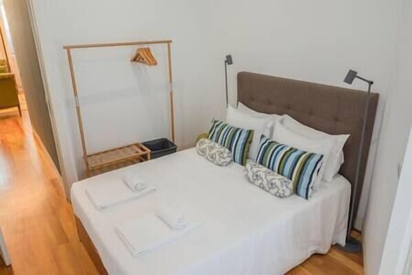Una cama bien hecha con sábanas blancas y almohadas a rayas, una mesita de noche con lámpara y un perchero de madera en la pared.
