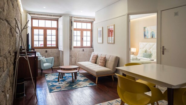 Ein gemütliches Wohnzimmer mit einer Couch, einem Couchtisch und einem Esstisch mit gelben Stühlen. Zwei Fenster mit Holzrahmen und ein Gemälde an der Wand sind zu sehen.